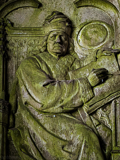 A weathered stone relief sculpture, heavily covered in green moss and lichen.

The sculpture depicts a scholarly figure, carved in profile and three-quarter view. He is wearing a distinct draped cap and thick, heavy robes with fur-lined or wide cuffs. 

He is shown actively working, holding an open book in his lap. His right hand holds a stylus poised against the page, while his left hand rests near the top of the manuscript.