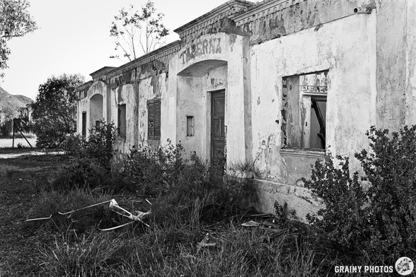 A dilapidated building with peeling paint and broken windows, surrounded by overgrown grass and scattered debris. The structure has a weathered, historic appearance, suggesting abandonment and decay.