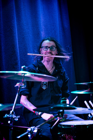 Auf dem Foto ist der Schlagzeuger der Band Lunar Fall - Max - zu sehen. Er sitzt hinter seinem Schlagzeug und blickt mit einem seiner Drumsticks zwischen den Zähnen zur Kamera.