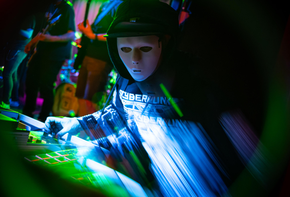 Ein Foto von B0N3, dem DJ und Scratcher der Band Lunar Fall. Er ist hinter seinem DJ-Pult abgebildet und trägt unter seinem schwarzen Hoodie und seiner Kappe eine weiße Maske, die sein Gesicht verbirgt. Das Bild ist ebenfalls mit einem künstlerischen Unschärfe-Effekt versehen.