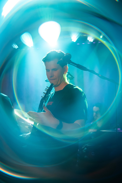 Das Foto zeigt Timo, den Gitarristen der Band Lunar Fall auf der Bühne mit seiner Gitarre in der Hand. Auch dieses Bild ist mit künstlerischen Reflexionen und Ringen aus Licht versehen. Im Hintergrund ist unscharf der Schlagzeuger Max hinter seinem Schlagzeug erkennbar.