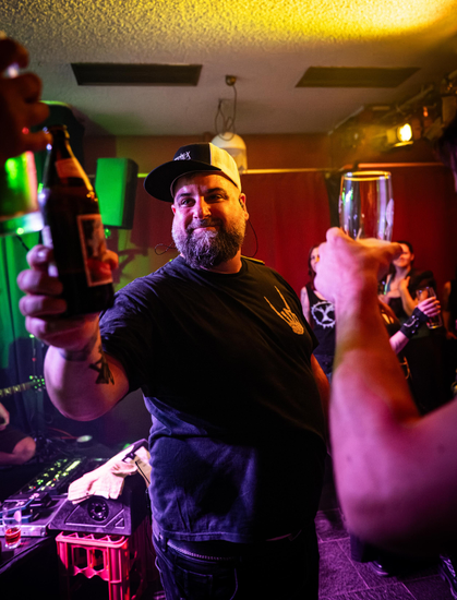 Auf dem Foto ist Babak der Sänger der Band Lunar Fall abgebildet, der vor der Bühne im Publikum steht und Personen im Publikum mit einer Flasche Bier zuprostet. Von links und rechts ragen Arme mit Getränkegläsern in Richtung Babak ins Bild und im Hintergrund sind weitere Personen aus dem Publikum zu sehen.