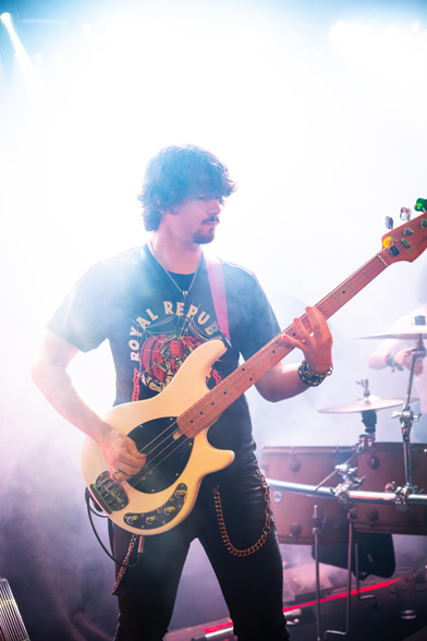 Das Foto zeigt Felix, den Bassisten der Band Lunar Fall, während er mit seinem Bass in der Hand umhüllt von weißem Nebel vor dem Schlagzeug auf der Bühne steht. Da der Nebel von hinten beleuchtet wird, scheint er hell zu strahlen.
