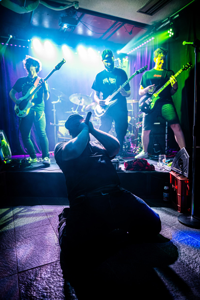Das Bild zeigt die Band Lunar Fall mit Ausnahme des DJs der Band auf beziehungsweise vor der Bühne. Die beiden Gitarristen sowie der Bassist stehen mit ihren jeweiligen Instrumenten am vorderen Bühnenrand, während der Sänger vor der Bühne nach hinten gelehnt auf dem Boden kniet, sich mit beiden Armen das Mikrofon vor das Gesicht hält und hinein singt. Der Schlagzeuger ist im Hintergrund zu sehen, jedoch wird er zum Großteil von seinem Schlagzeug und von einem der Gitarristen verdeckt.