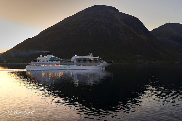 This is a colour landscape and seascape photo photo of a medium sized cruise ship underway through a wide sea fjord at sunset with a mountain ridge in the background. Ullsfjorden, Norway (2018).

The calm fjord water rises from the base of the photo upwards to the halfway up level. The water is disturbed only by the smallest of ripples. Above the water is a barren, brown mountain ridge, with the mountain's summit being in the centre and top of the image and sloping downwards gently towards the left. To the right there is a further more distant mountain peak. The closer mountain casts a shadow in the water below, extending to the base of the photo. Where the shadow touches the water: the water is jet black, outside the shadow the water reflects the clear and cloudless sky. The setting sun (near midnight sun) is out of view to the left of the photo and is casting a golden glow over the water in the lower left corner. From the image's centre and stretching to the left is our cruise ship, underway from the left to the right, viewed in profile and occupying half the width of the photo. The ship is all white, has a sharp, pointed bow and her lights are casting an orange and golden glow on the water below. The vessels streamlined funnel is trailing a thin wisp of white diesel exhaust smoke. 

The cruise ship is the 210m long MV Seabourn Ovation steaming through the Ullsfjorden in Norway's arctic north. 