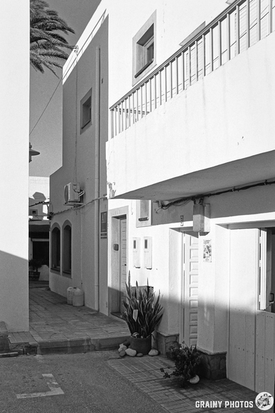 A narrow alleyway between two white buildings, featuring a balcony and tropical plants. The scene is captured in black and white, enhancing the architectural details and shadows of the surroundings.