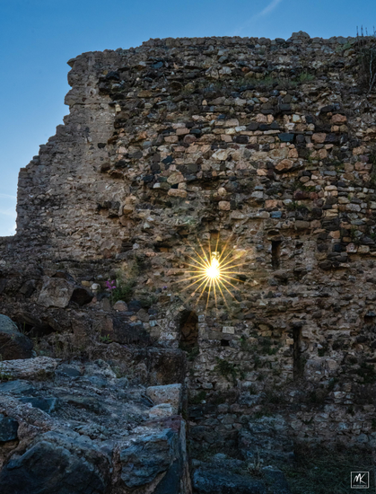 Color photo of a rayed burst of light from the setting sun passing through a narrow vertical window or loop hole in the ruined stone wall of a medieval castle. 