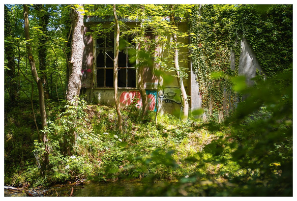 Ein Gebäude steht dicht an Bäumen und einem Bach im Wald. Einige Grafitti-Schmierereien sind zu sehen. Das Gebäude wirkt verlassen und verfallen