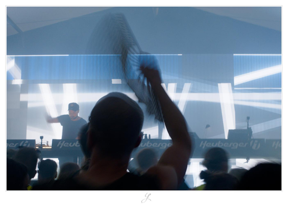 The view is towards the stage on which the DJ stands behind the mixing consoles, raising his right arm, blurred by the movement. Directly in front of the photographer, someone is fanning himself with a fan and the people around him. The people are visible as dark silhouettes against a blue-and-white animation on a large screen behind the DJ. The air is filled with artificial fog.

AI disclaimer: Using my work, its meta data, written or derived description to create media with or train AI based systems is prohibited.