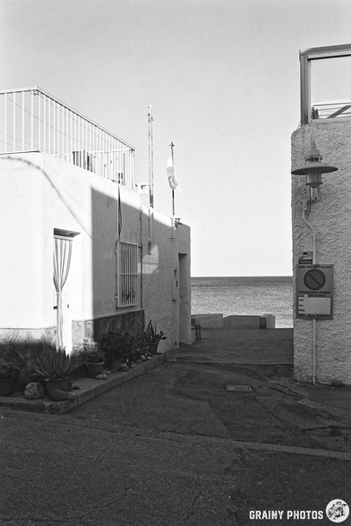 A narrow passageway between white-washed buildings leads to the calm sea, with a flag waving gently in the breeze. Potted plants adorn the walkway, and a street lamp stands quietly as the sun casts long shadows in this serene coastal setting.