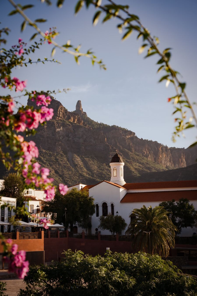 Pink flowers in the foreground, a white church and a few white houses in the main image, and a single rock peaking above the mountains in the background