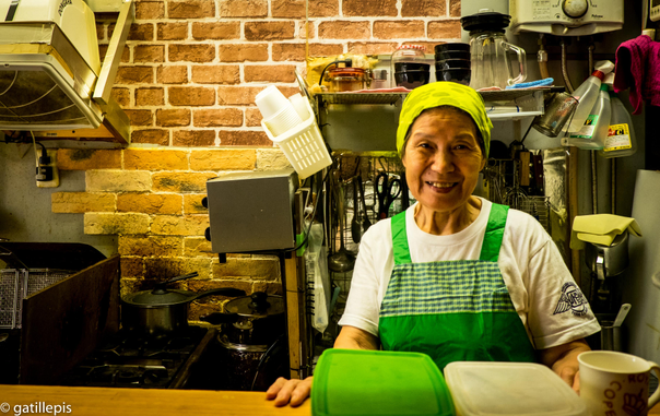 Dona mirant a càmera. Aquesta dona tenia un petit bar on feia esmorzars tradicionals japonesos (truita, peix, sopa de miso, etc.). Va aprendre l'ofici de cuinar treballant en una base americana i per això, també, parlava força bé l'ànglès. Al cap d'uns anys hi vag tornar per saludar-la i el bar havia canviat de propietari i d'esil.