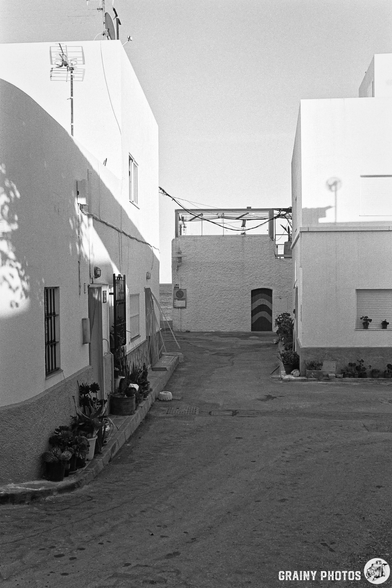 A narrow street lined with white buildings, featuring a small entrance in the background. Potted plants adorn the edges, and a clear sky above suggests a sunny day. The scene is captured in black and white, highlighting the architectural details.