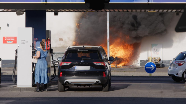 Eine Frau in einem graublauen Kleid tankt ihr Auto auf, während im Hintergrund, ca. 10 Meter weiter, ein Brand lodert