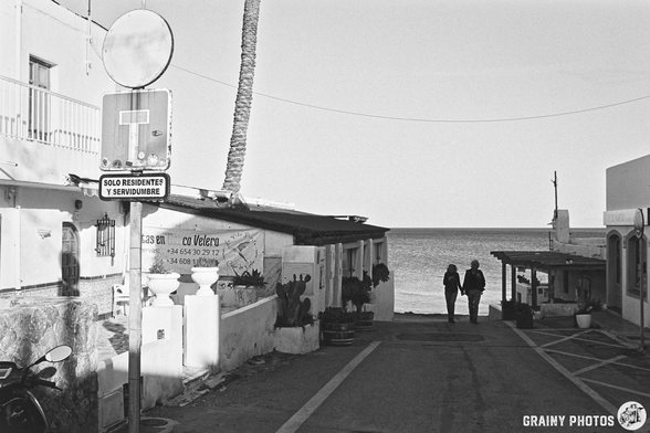 A narrow street lined with white buildings, featuring a small entrance in the background. Potted plants adorn the edges, and a clear sky above indicating a sunny day. The scene is captured in black and white, highlighting the architectural details.