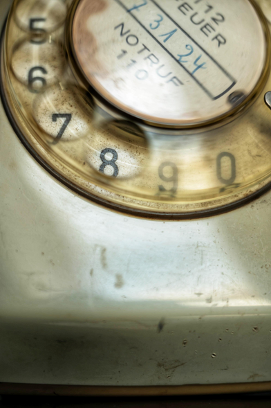 Die Nahaufnahme zeigt die Wählscheibe eines historischen, elfenbeinfarbenen Telefons aus einer leicht schrägen Perspektive von oben. Im Zentrum der gläsernen Wählscheibe befindet sich ein runder, weißer Papiereinsatz, auf dem oben der Name „EUER“ und darunter handschriftlich die Telefonnummer „73 1 24“ eingetragen ist. Darunter steht deutlich der gedruckte Schriftzug „NOTRUF 110“. Die Wählscheibe selbst besteht aus transparentem Kunststoff mit kreisförmigen Vertiefungen für die Finger, unter denen die schwarzen Ziffern auf hellem Grund erscheinen; im Bildausschnitt sind die Zahlen 5 bis 0 deutlich erkennbar wobei im ersten Kreis die 8 erkennbar ist. Das Gehäuse des Telefons weist eine glatte, matte Oberfläche mit leichten Gebrauchsspuren und feinen Kratzern auf, die den antiken Charakter unterstreichen. Die warme Beleuchtung erzeugt sanfte Reflexionen auf dem Glas und den Rundungen des Geräts, während der untere Teil des Bildes in eine leichte Unschärfe verläuft.