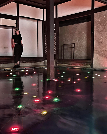 Eine Kunstinstallation: auf einer Wasserfläche sind sehr viele bunte, neonfarben leuchtende Ziffern im Wasser zu sehen. Am hinteren Rand steht vor einer hellen Wand eine dunkelhaarige Person, die sich den Raum ansieht. Der Raum ist durch schmale dunklere Elemente vertikal und horizontal gegliedert.