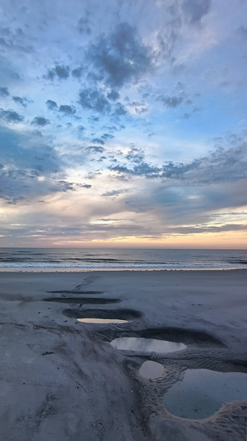 Serene beach at dusk with gentle waves and a tranquil sky, featuring scattered clouds. Puddles in the foreground reflect the soft pastel colors.
