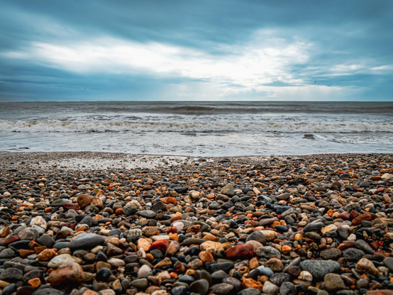 Wide shot of a pebble beach covered in smooth, multicolored stones in shades of grey, brown, orange, red, and cream. Calm white waves roll onto the shoreline while the grey sea leads out to the horizon under a dramatic, cloudy blue sky.