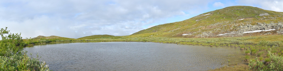 A panoramic photo of a pond on a mountain. The mountain has low growing plants on it, and continues rising to the right of the image. The sky is blue and filled with white clouds.