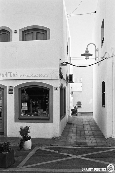 A narrow street between white buildings featuring a shop called "Maneras" with a large window display. The scene is illuminated by a street lamp, and there's a potted plant by the entrance, suggesting a quaint, coastal atmosphere.