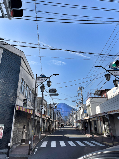 Eine Straßenflucht in einer nur niedrig bebauten japanischen Ortschaft. Am Ende ist der Berg Fuji zu sehen. Im Vordergrund ein abbiegendes Auto im Anschnitt und links oben eine Ampel im Anschnitt. 