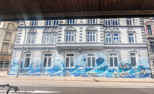 Eine helle Altbaufassade eines mehrstöckigen Gründerzeit-Gebaudes. Auf der gesamten Breite des Erdgeschosses, über sechs Fenster hinweg sind mehrere Wellen im Stil Hokusais "Großer Welle von Kanagawa" modern umgesetzt. Der Blick ist aus dem Tunnel kommend, noch mit etwas  Tunneldecke am oberen Rand im September 2023 aufgenommen worden. 