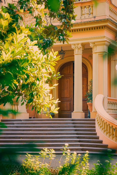This is a photograph of a grand, classical-style building doorway. There is a wooden double door with carvings which is set in an arched frame. It is supported by ornate, cream-coloured columns. There are also wide stone steps that lead up to the entrance. The door and the stepd are flanked by hreenery with sunlight filtering through leaves. Oh, there's also a black lantern which is hanging above the door and a potted plant sits on the right.