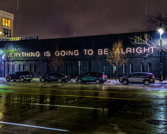 Night view of the Museum of Contemporary Art Detroit taken from across the street on a rainy night. Large neon text reads "EVERYTHING IS GOING TO BE ALRIGHT" across a long black wall. Four vehicles are parked along the curb on a wet, reflective asphalt street.
