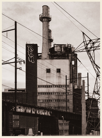 Sepia-toned photograph of an industrial building with smokestacks and power transmission towers. A tall black structure bears a large “REW” graffiti tag, while power lines crisscross the scene and an elevated rail bridge with additional graffiti runs across the foreground.