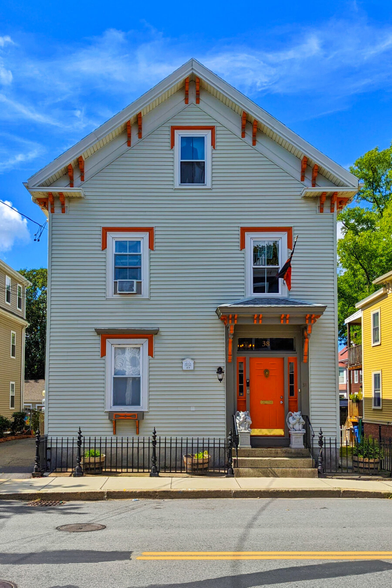 A photo of a striking Victorian house under a blue sky with wispy clouds taken from across an asphalt street with yellow road markings. The three-story house has light gray horizontal siding and striking bright orange trim. A steep gable roof is decorated with orange brackets and the tall narrow windows also have bright orange hoods. The vibrant orange front door is flanked by two small gargoyle statues while a low black iron fence separates the house from the sidewalk.