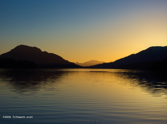 Beautiful cloudless blue sky with diminishing light, over the loch which is bounded by silhouetted mountains.  The setting sun is creating a lovely warm glow, which nearly obscures one distant mountain in the centre of the photo.
