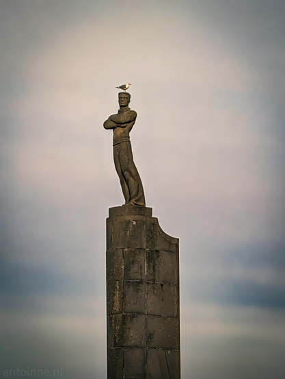 A tall, vertical, dark stone cylindrical column with a statue of a man. He has his arms crossed, is leaning slightly forward, and has a seagull sitting on his head. The background is a vast, open sky filled with soft, gray clouds.