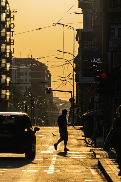 A photograph of via Saffi at sunset, bathed in golden light. In the middle, there is a silhouetted man in casual clothes and he's walking across the road. There is also a silhouetted car on the left and a bus stop on the right. Some overhead tram lines, signs and traffic lights are visible, with the sky glowing orange. The street looks wet and is reflecting the golden light. (It's not wet, I don't actually know why it's so reflective!)