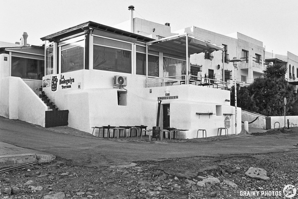 A black and white photograph of a modern restaurant with large windows, located on a hillside. The building features white walls, outdoor seating, and a surrounding rocky area, typical of a coastal town.