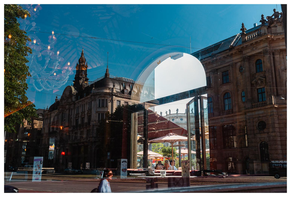 Blick durch eine spiegelnde Fensterscheibe in und durch ein Cafe und wieder hinaus. In der Spiegelung der Scheibe sind große Häuser, Passanten, Himmel zu sehen.