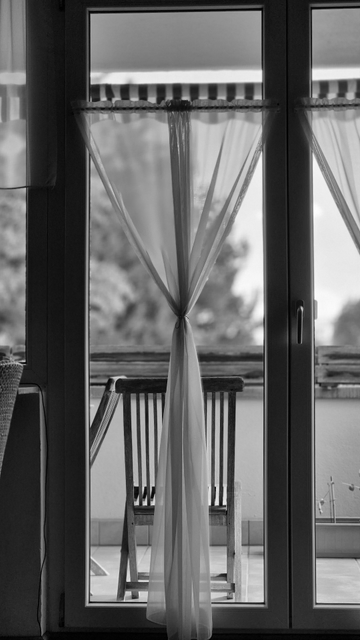 Ein rechteckiges Hochformat-Foto in schwarzweiß. Das Foto zeigt eine häusliche Szene. Von innen ist der Raum so gestaltet, dass der Blick durch die geschlossenen, bodentiefen verglasten Türen auf den Balkon führt. Im Bildzentrum befindet sich der linke Türflügel, während der rechte Türflügel lediglich einen kleinen Teil des Bildes einnimmt. Vor der Glasscheibe ist ein Store angebracht, der in seiner Breite mit einem Band zusammengerafft ist. Dadurch wird ein freier Blick nach draußen ermöglicht. Vor der Tür befindet sich ein Gartenstuhl aus Holz, der mit einer Rückenlehne aus senkrecht stehenden Holzstäben ausgestattet ist. In der Ferne sind hinter der geschlossenen Balkonbrüstung unscharf Bäume zu erkennen. 