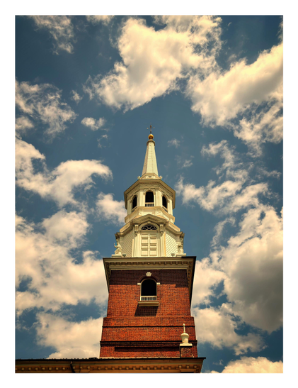 Photo taken from street level looking up at the sunlit bell tower of a small church, framed by cottony white clouds in a blue sky. The four-sided lower part of the tower is made of warm red brick; an arched window with a low white guardrail is set in the center of the facade. The upper sections are eight-sided (with three sides visible) and made of white painted wood; here, an arched window with a covered main panel and a triangular pediment sits between two small statues, while above it three smaller arched windows look out from their respective sides. At the top is a white wooden spire tapering to a golden ball and a metal cross.