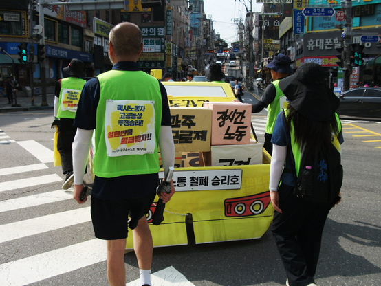 역곡역에서 출발하는 행진대오 선두에서 택시 모형을 끌고 가는 택시노동자들. 택시 모형 트렁크에는 “간주근로제”, “장시간 노동” 따위가 있다.