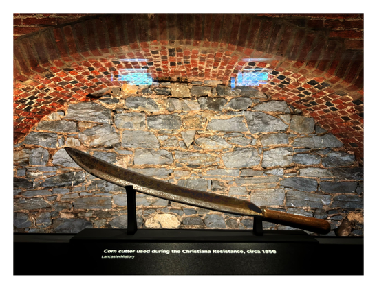 A rusted corn cutter — a farm implement with a wooden handle and long curved blade — is displayed in front of a wall of stacked stones under an arch of bricks. A label below the blade reads “Corn cutter used during the Christiana Resistance, circa 1850.” Photo taken at the Thaddeus Stevens & Lydia Hamilton Smith Center in Lancaster, Pennsylvania.