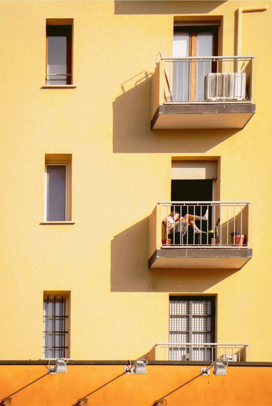 This is a photograph of a yellow  building with three rectangular windows and three small balconies. The middle balcony has a person with light brown hair leaning against the railing, wearing a white shirt. At the bottom there is an orange wall with lights which cast shadows. To the left of the balconies are three vertical windows