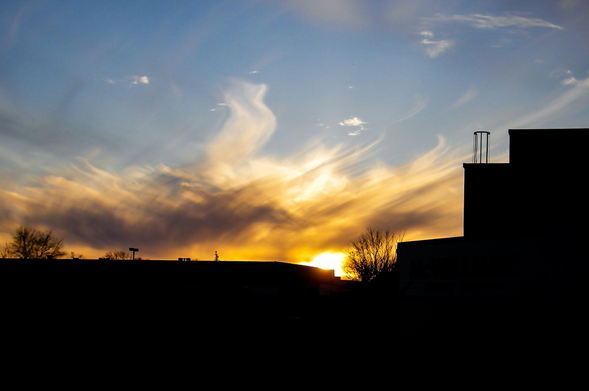 Soft dramatic clouds allow the setting suns yellow glow to shine through them giving the impression of flames. The silhouette of building rooftops can be seen along the bottom. 
