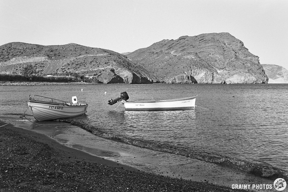 A black and white photograph of a modern restaurant with large windows, located on a hillside. The building features white walls, outdoor seating, and a surrounding rocky area, typical of a coastal town.