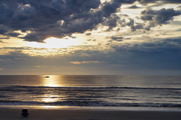 A serene beach at sunrise with dramatic clouds. Sunlight reflects on calm waves, and a lone boat is visible on the horizon. A chair rests on the sand.