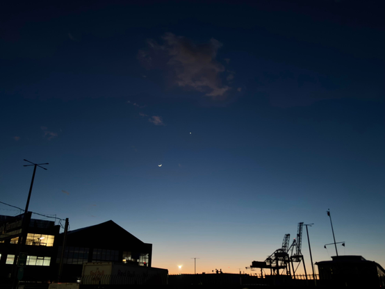 The Brooklyn waterfront just after sunset. The shadowy foreground features silhouettes of shipping cranes and warehouses with lit windows. Above is a wide sky, deepening blue and transitioning to gold at the horizon, mostly clear except for a puff of orange cloud at the center top of the photo. Below the cloud is a tiny crescent moon, and the bright point of Venus to the moon’s upper right.