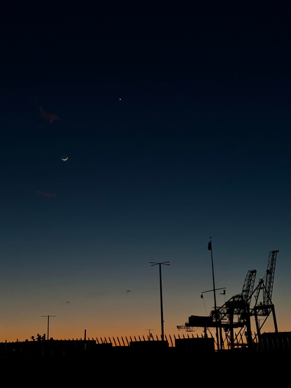 Photo of the same scene with a closer view of the silhouettes of shipping cranes and the moon and Venus above. Far below the moon, just where the sky begins to shift from blue to orange-gold, is the planet Mercury, showing as a very faint point of white light.