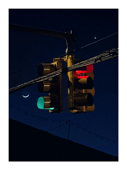 Closeup view of a three-way traffic signal suspended among power lines against a dark evening sky. The signal is flanked by a small crescent moon on its lower left and Venus as a bright point on its upper right. On its two visible sides, the green light is activated on the left and the red is activated on the right. Below is the oblique silhouette of a warehouse rooftop.