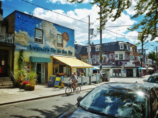 A cyclist rides past a restaurant called "Wanda's Pie In The Sky" with a mural depicting a blue sky with large white clouds. A blue sky with white clouds is shown along with other buildings in the background as well as cars parked along the street.