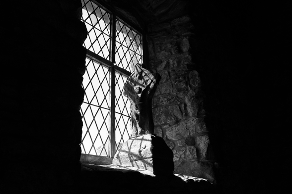 In einer kleinen Nische mit vergittertem Fenster steht eine kleine Skulptur mit einer in Stein gehauenen Figur, die die Hände über den Kopf hebt. Licht fällt durch das Fenster und der Rest des Bildes ist dunkel, fast schwarz.

In a small alcove with a barred window stands a small sculpture featuring a figure carved from stone, raising its hands above its head. Light streams through the window, whilst the rest of the scene is dark, almost black.