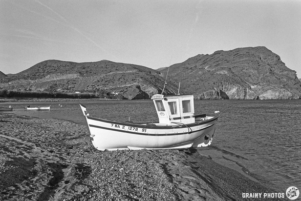 A black and white photograph of a small fishing boat resting on a pebbly beach, with rocky hills in the background and calm water in the foreground, capturing a serene coastal landscape.